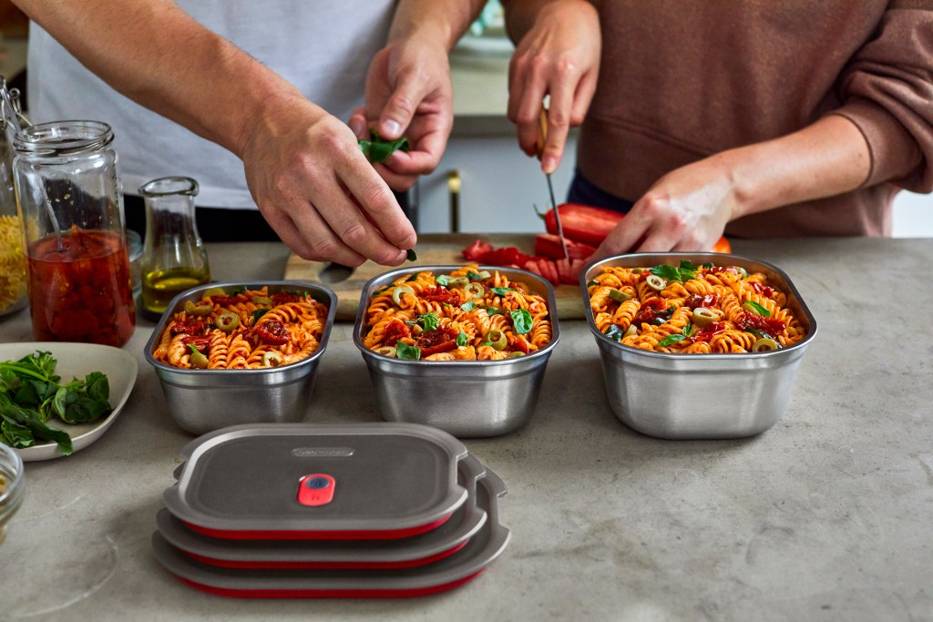 Two people preparing food in a kitchen, with colorful pasta dishes in stainless steel meal prep containers on the counter.