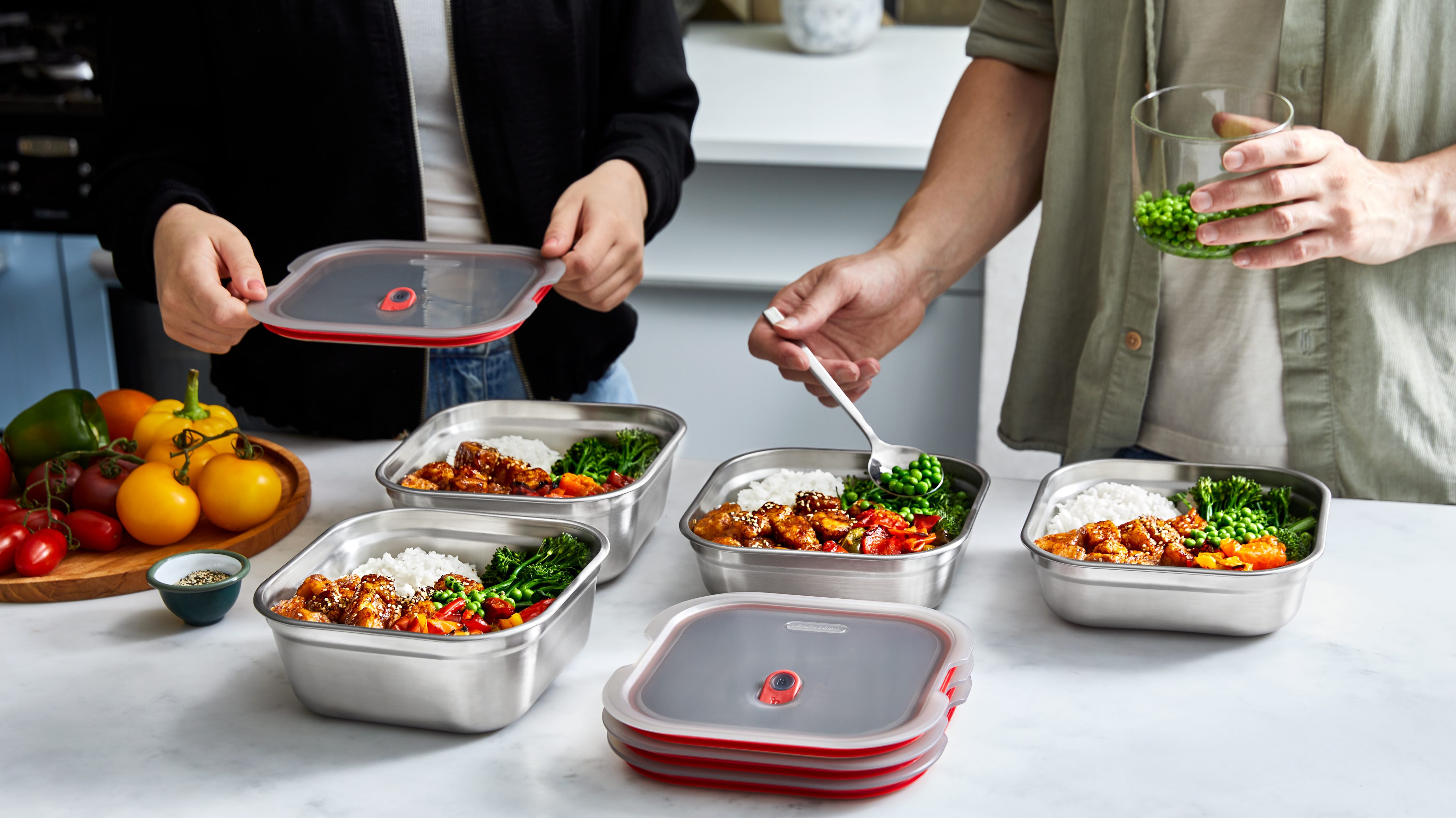 Two people preparing meal prep containers on a countertop, with colorful vegetables and food in stainless steel boxes and red lids.