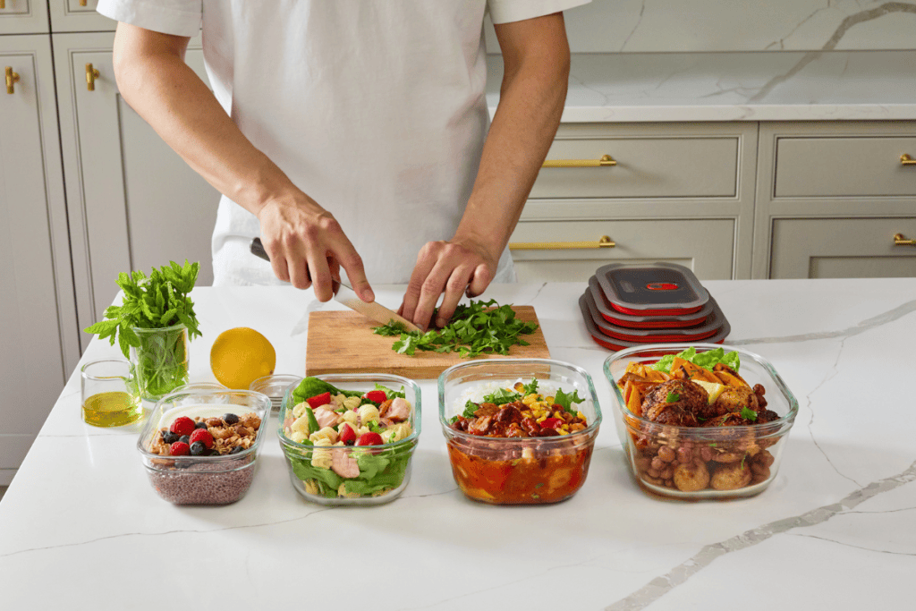 A person chopping fresh herbs on a wooden cutting board in a modern kitchen, with various meal prep containers filled with healthy food, including salads and cooked dishes, on a white countertop.